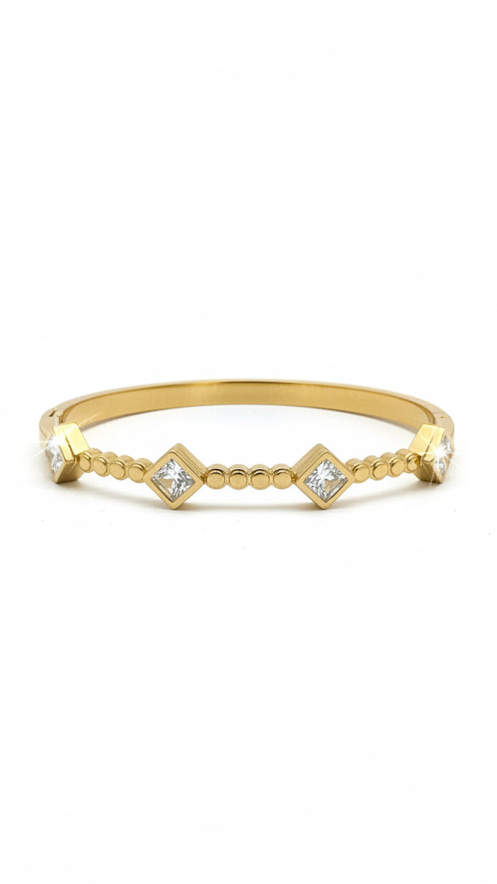 A gold-plated bangle bracelet with diamond-shaped crystal accents, displayed on its white branded retail card inside a clear plastic sleeve on a dark desk.